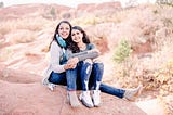 A half-black mother with mid-length dark brown hair, wearing a grey sweater and blue jeans, hugs her teenage daughter, a half-black girl with long brown hair, wearing a long sleeve gray shirt and blue jeans. They are sitting atop a rock at Garden of the Gods park in Colorado.