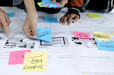 Photo of a table with printed papers, post-it notes and people interacting with the notes in a workshop.