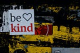 “Be Kind” sign on wall with black and yellow graffiti