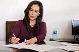 An attractive dark haired lady sits writing at an office desk.