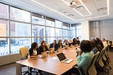 Several women of color and a couple of men sit at a large light brown conference room table with laptops in front of them. The conference room has large windows. The atmosphere appears fun and lighthearted.