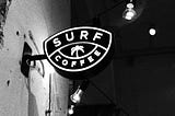 A sign on a building, that says Surf and Coffee. There is also an image of a palm tree. The image is black and white, possible also a night time scene.
