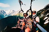 A man and woman are shown in harness gliding. Snow capped mountains are shown in the background