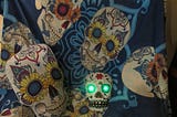 A photo of multiple layers of “day of the dead” skulls of various sizes — gayly decorated with colorful flowers and geometric designs — the center skull has erily gleeming green eyes.