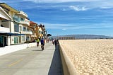 a pedestrian-only street next to the beach, with houses on one side and sand on the other. it’s a blue-sky day.