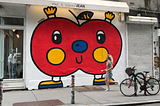 Street art — a red apple by the artist Sonni is raising its arms and rejoicing, on a white wall in NYC Bowery. A white woman in a sporty dress runs past the apple in a hurry.