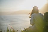 We see a woman sitting on the shore, looking toward the ocean and mountains. We see her from the back. The sun shines.