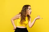 Young woman against a bright yellow background, pointing to someone out of view and looking shocked.