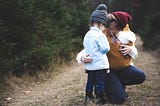 A woman baby wearing an infant and squatting down to talk to a toddler.