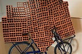 Dozens of large red clay mud bricks loaded on a black metal bicycle in an art installation of 21st century art. The bricks seemingly overwhelm the bike, which is still, however, standing.