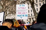 Photograph of a sign at Ban conversion therapy for all — The Trans Rights Protest London, April 2022 that reads “If it’s utterly abhorrent for LGB, it must be for T” in the transgender flag colors.