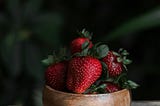 The central image is of a wooden bowl filled with ripe red strawberries sitting on a wooden table with a newspaper sheet. dark background.