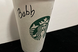 A Starbucks cup sits on a black counter. The name Bob (misspelled as “Bobb”) is written on it in black marker.