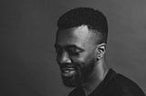 A black and white headshot of Daniel, a Black man with a fade who is smiling slightly in a black t-shirt