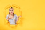 Grumpy looking woman, long blonde hair in plaits, red lipstick, pushing through yellow paper. Her hands are palm-up and pointing to her left as if she is explaining something to an idiot.
