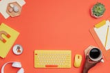 Office supplies, a yellow typewriter, yellow computer mouse, white headphones, green succulent plant, orange and yellow notepad, and hand of Black person holding a coffee mug with black coffee on an orange desk.