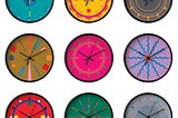 Nine clock with black hands and black edges are pictured, arranged in an evenly spaced grid on a white background. Each clock face has a different geometric, colorful design, on backgrounds that are turquoise, yellow, grey, ochre, hot magenta, lavender, red, green and grey.