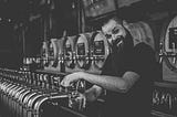 A person at a pub bar fills a pint glass, looking at the camera. There are several casks in the background.