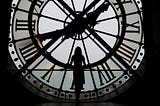 The silhouette of a tall woman stands at a large portrait window facing a clock