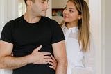 Man and woman look at each other, he is standing slightly to the front, wearing a black top, with his hand holding his partner’s hand. She stands slightly behind wearing a white dress. They look at each other with affection.