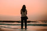 A woman stands alone at the edge of the ocean at sunrise, looking out across the water beneath a soft pink sky, symbolizing reflection, resilience, and new beginnings.