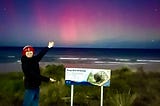 The author wearing a red hat standing next to a sign. Behind him, the ocean waves are visible with a bright red aurora in the night sky.