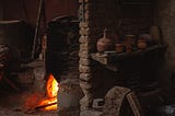 A firing kiln glowing red hot behind a brick wall holding a shelf with clay pots.