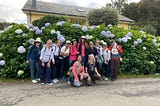 A group of thirteen women pose for the camera on the street in front of a tall hydrangea bush with large blue and white flowers.