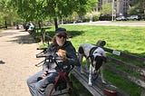 Photo of an older woman sitting next to a park bench with a dog.