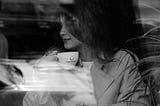 Woman sitting alone at a café window, holding a cup of coffee, seen through glass.