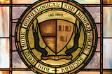 A stained glass window depicting the seal of the New York Genealogical and Biographical Society. The seal is divided in half, consisting of a rising sun over water on the left and three open books on the right.