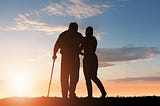 Two people in silhouette walking toward sunset, one is using cane in left hand and slightly leaning on the other person