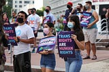 Masked protestors of different genders and races stand with signs that read “Trans Rights are Human Rights” and “#LetKidsPlay Equality Florida”