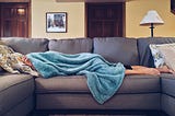 Person lying on gray sofa, a fuzzy, blue blanket covering their whole body minus their calves and bare feet