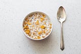 A bowl of cereal with milk and a silver spoon rest on a table top