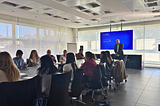 A professional corporate training session for Touchpoint Strategies by Knowcrunch is taking place in a modern conference room, where a presenter stands near a screen displaying a welcome message while participants engage in discussion around a large table.
