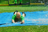 It’s a sunny, hot summer day, and a bald man is sliding face-forward toward the camera on a homemade slip-n-slide in a beautiful, green field. He is in a green shirt and you can see his while legs and black shoes as he’s almost to the end of the watered down, blue tarp. To young men in are in the background watching him (with matching green shirts and dark shorts) waiting their turn to “slide.”