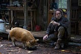 Robin (played by Nicolas Cage) sits on the porch of his cabin holding a piece of pie. His brown pig is next to him and eating leftovers out of a skillet.