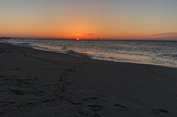 The sun rises out of the ocean, it looks like a pink peach appearing out of the dark-velvety-blue, calm ocean.