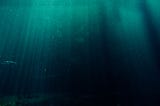 fundo do mar, com raios de luz atravessando a imagem em linhas verticais. As águas fazem um degradê de verde azulado até completamente preto.