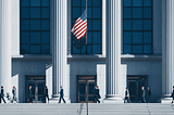 Professionals walking up the steps of a federal government building with tall columns and an American flag, representing the U.S. contracting and business sector. Discover how small federal contractors can create lasting growth through operational flywheels that compound performance, trust, and capability.