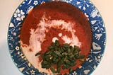 Una sopa roja en un cuenco azul. Hay una espiral blanca de leche de coco y una montañita de semillas de calabaza.