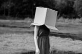 A young, barefoot woman stands alone in a grassy field with a cardboard box over her head.