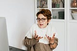 Woman with glasses and white and brown sweater shrugging to show how little she cares what men think about her.