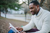 man with white shirt sitting in nature reading a book