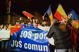 Enthusiastic protesters at night in Cluj-Napoca, Romania, waving Romanian flags and holding a banner with the message “JOS ponta JOS comunismul” (DOWN with Ponta DOWN with communism). The atmosphere is vibrant with citizens actively participating in the democratic process, calling for political change.