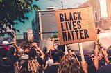 Protestors crowd a street, facing away from the camera. Facing the camera is a cardboard sign saying Black Lives Matter.