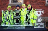 Two men and two women stand behind a 40" wide ice sculpture that reads “WOW MOM”. A 20" tall trophy made of ice that reads “Carver’s Choice 2022” rests next to the sculpture. All four people are smiling.