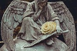 A statue of a seated angel in a cemetery holds a single white rose.