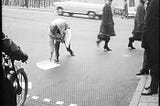 A black and white photo of a man putting a3 pieces of paper on the floor on a busy street with pedestrians walking.